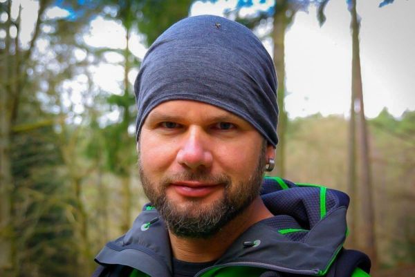 Mann mit grauem Kopftuch und schwarzer Jacke im Wald