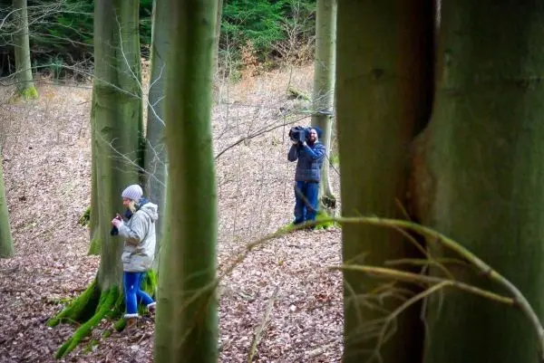 Zwei Personen stehen zwischen Bäumen im Wald, eine nutzt ein Smartphone, der andere hält eine Kamera