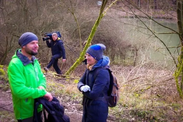 Zwei Personen unterhalten sich am Ufer eines Gewässers, während eine dritte Person mit Kamera vorbeigeht