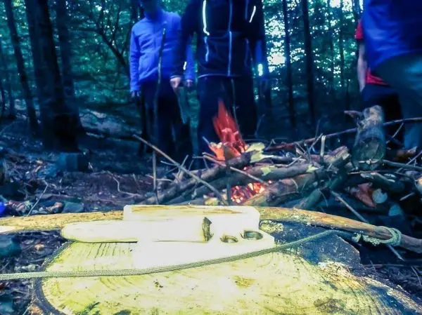Holzstück mit Schnur und Werkzeug liegt vor einem kleinen Feuer im Wald