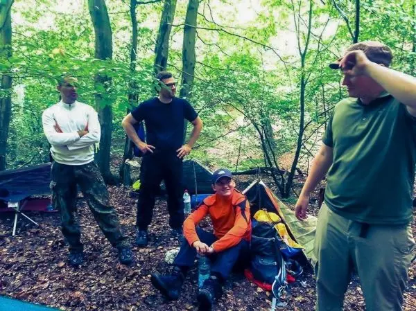 Vier Personen stehen und sitzen in einem Wald neben einem Zelt
