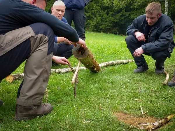 Drei Personen arbeiten an einer Holzhebel-Konstruktion mit einem Baumstamm und einem Stock