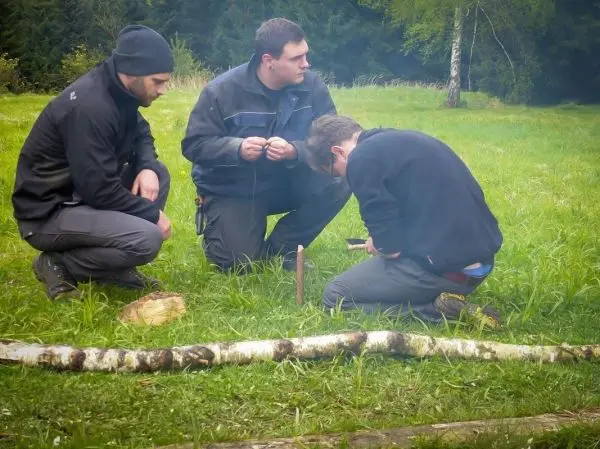 Drei Personen arbeiten auf einer Wiese an einer Holzarbeit mit einem Stock