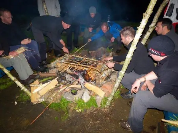 Grillrost aus Holz über einem offenen Feuer, mehrere Personen bereiten Essen vor