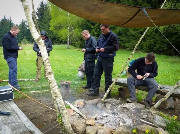 Gruppe bearbeitet Holzstücke am Lagerfeuer unter einem Tarp