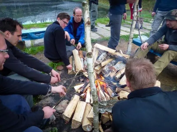 Gruppe sitzt um ein Feuer und röstet Essen auf Spießen beim Survival Training