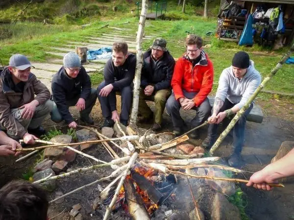 Gruppe sitzt um ein Feuer und röstet Stöcke beim Survival Training
