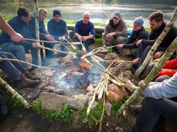 Gruppe sitzt um ein Feuer und röstet Stöcke beim Survival Training
