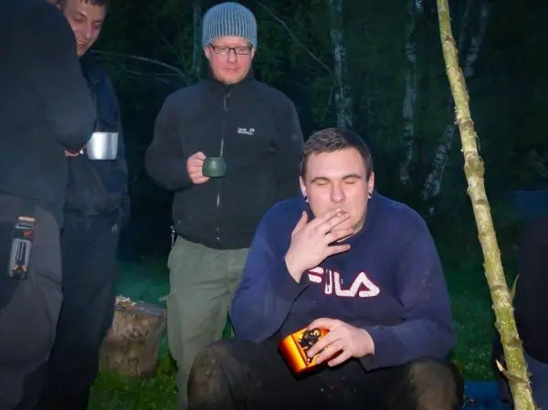 Gruppe steht um ein kleines Feuer, einer raucht und hält eine Dose