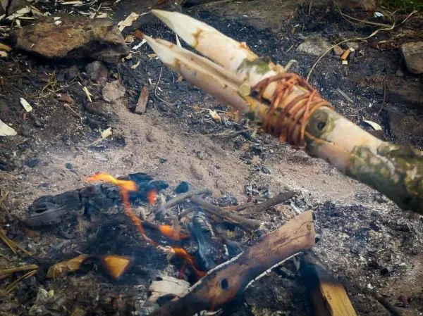 Kleine Flamme glimmt im Bodenfeuer, improvisierte Speerspitze aus Holz sichtbar