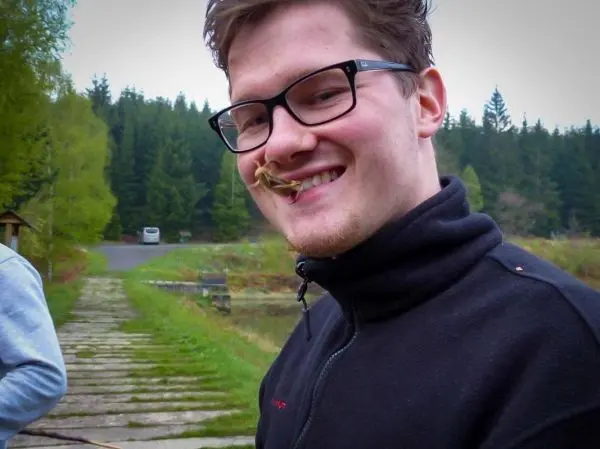 Lächelnde Person mit Brille hält einen Zweig im Mund, Hintergrund mit Bäumen