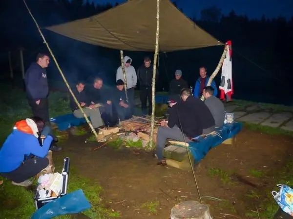 Lagerfeuer brennt unter einem Tarp, umringt von mehreren Personen auf Unterlagen