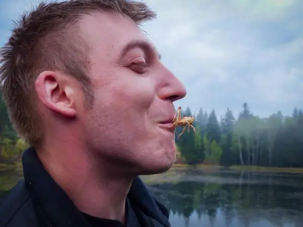 Mann hält eine Heuschrecke zwischen den Lippen am Ufer eines Gewässers