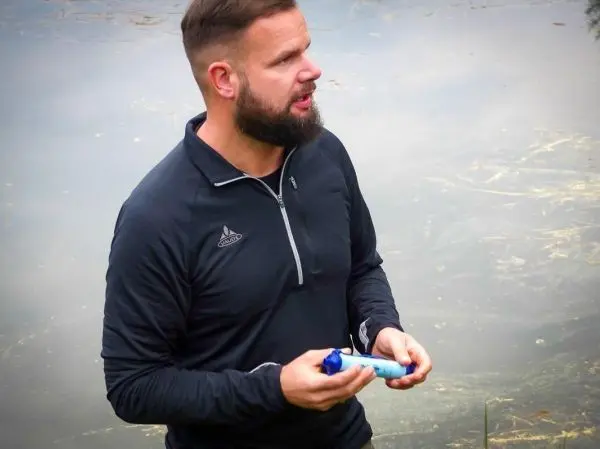 Mann hält einen blauen Wasserfilter in der Hand, steht am Ufer eines Gewässers