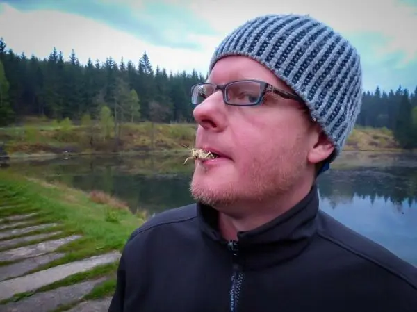 Mann mit Mütze steht am Ufer eines Gewässers und hält einen Grashalm im Mund