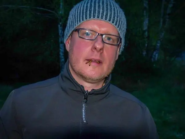 Mann mit Mütze und Brille steht im Freien, Holzstück im Mund