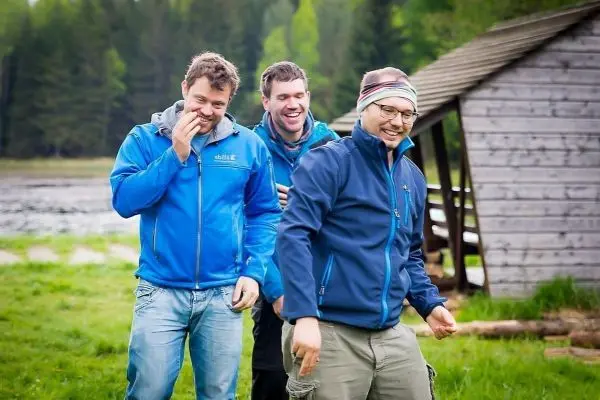 Drei Männer in blauen Jacken stehen lachend auf einer Wiese neben einer Holzhütte