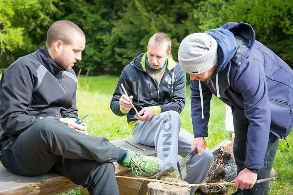 Drei Personen arbeiten an einem Holzstück mit einem Messer auf einer Holzbank