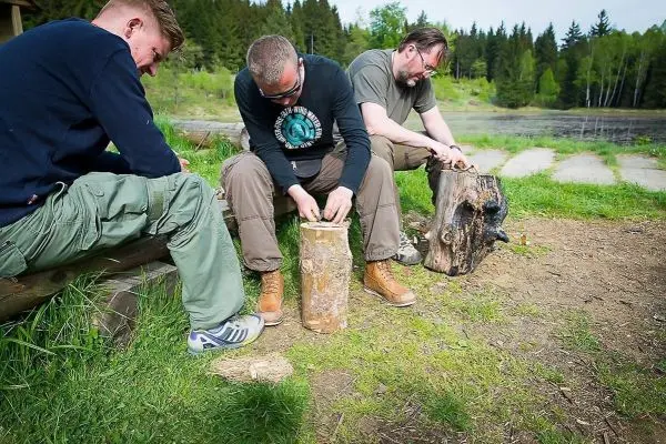 Drei Personen bearbeiten Holzstücke mit Messern am Ufer eines Gewässers