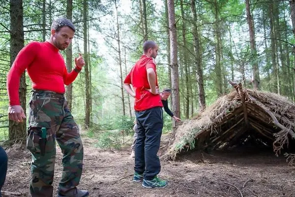 Drei Personen betrachten eine selbstgebaute Tarp-Notunterkunft im Wald