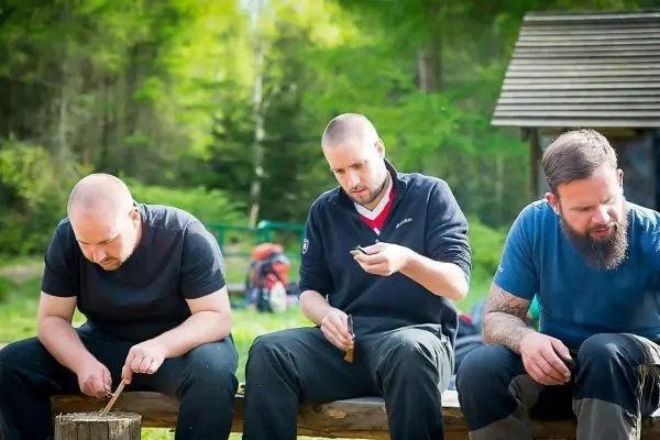 Drei Personen schnitzen Holz auf einer Bank im Freien