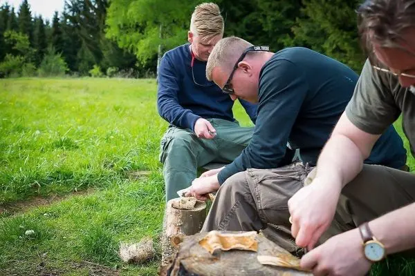 Drei Personen schnitzen Holzstücke auf Baumstämmen im Freien