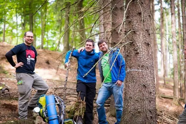 Drei Personen stehen neben einem Baum im Wald, Ausrüstung liegt am Boden