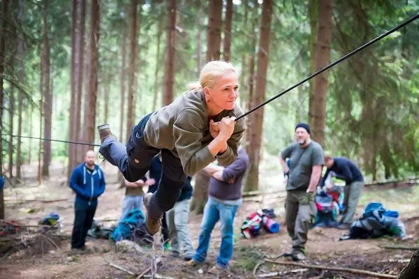 Frau überquert selbstgebaute Seilbrücke im Wald, Zuschauer im Hintergrund