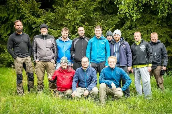 Gruppe von zehn Personen in Outdoor-Bekleidung steht auf einer Wiese vor Bäumen