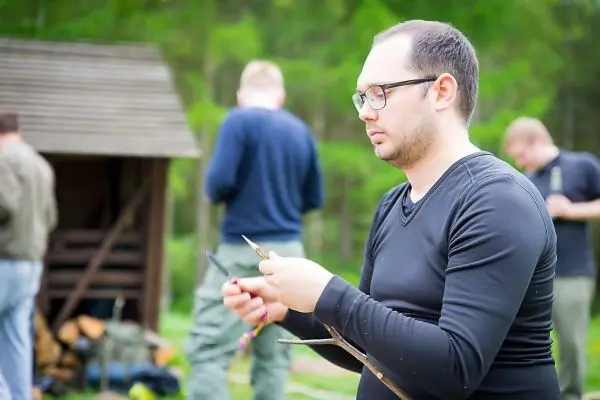 Mann bearbeitet einen Stock mit einem Messer, im Hintergrund eine Holzstruktur