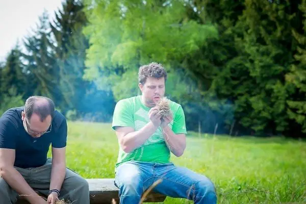 Mann bläst in Zundernest, um Glut zu erzeugen, während ein anderer sitzt