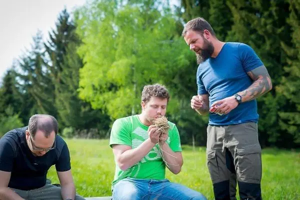 Mann formt Zundernest aus Gras, während zwei weitere Männer beobachten