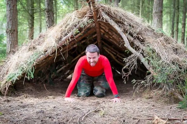 Mann kriecht aus einer selbstgebauten Laubhütte im Wald