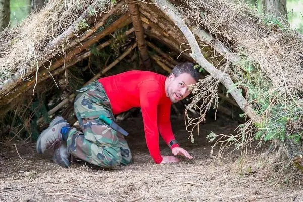 Mann kriecht in eine selbstgebaute Tarp-Notunterkunft aus Ästen und Zweigen
