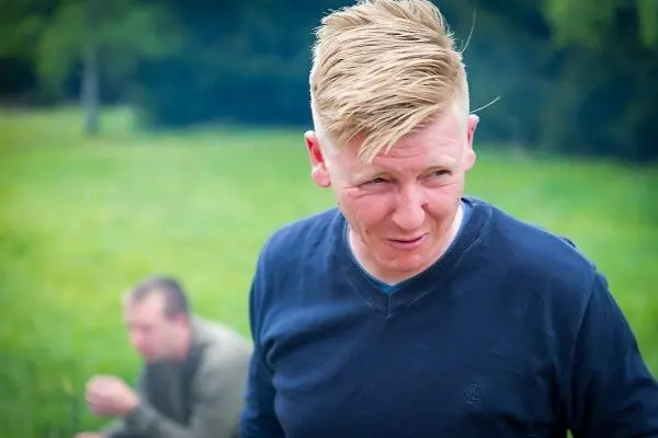 Mann mit kurzem, blondem Haar steht in einer Wiese, im Hintergrund sitzt eine weitere Person