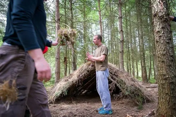 Mann mit Zunder steht vor einer selbstgebauten Tarp-Notunterkunft im Wald