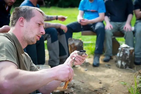 Mann schnitzt mit einem Messer an einem Holzstück, während andere im Hintergrund sitzen