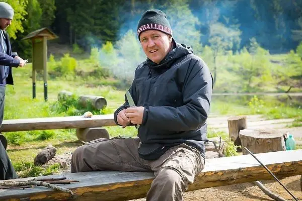 Mann sitzt auf Holzbank und schnitzt mit einem Messer an einem Stock