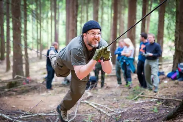 Mann überquert selbstgebaute Seilbrücke im Wald, andere Teilnehmer beobachten
