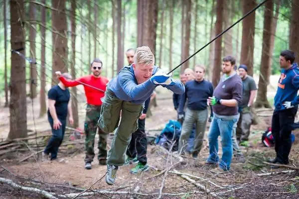 Mann überquert selbstgebaute Seilbrücke im Wald, Zuschauer beobachten