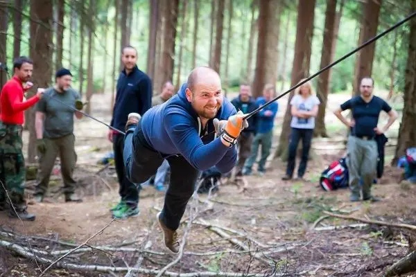 Mann überquert selbstgebaute Seilbrücke im Wald, Zuschauer beobachten
