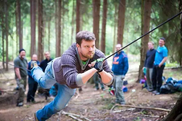 Mann überquert selbstgebaute Seilbrücke im Wald, Zuschauer im Hintergrund