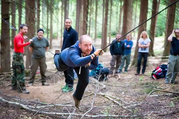 Mann überquert selbstgebaute Seilbrücke zwischen Bäumen, Zuschauer im Hintergrund