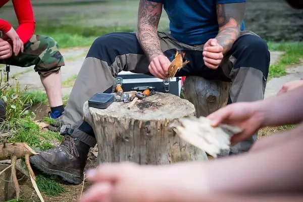 Messer schneidet Zunder auf einem Baumstumpf, während Personen im Hintergrund sitzen