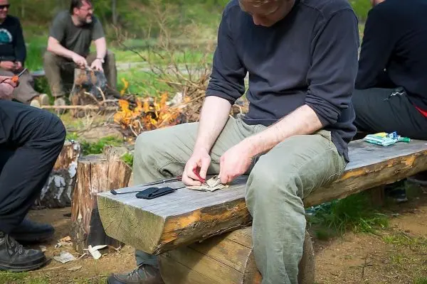 Messer schneidet Zunder auf einer Holzbank, im Hintergrund brennt ein Feuer