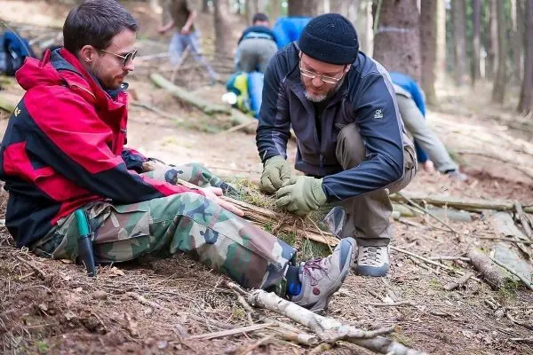 Person bindet Äste zusammen, Person sitzt daneben im Wald