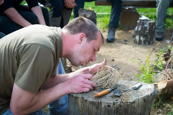 Person bläst in ein Nest aus Gras auf einem Baumstumpf beim Überlebenstraining