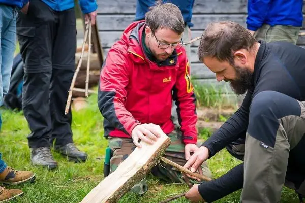Zwei Personen bearbeiten einen Holzstamm mit Stöcken beim Überlebenstraining