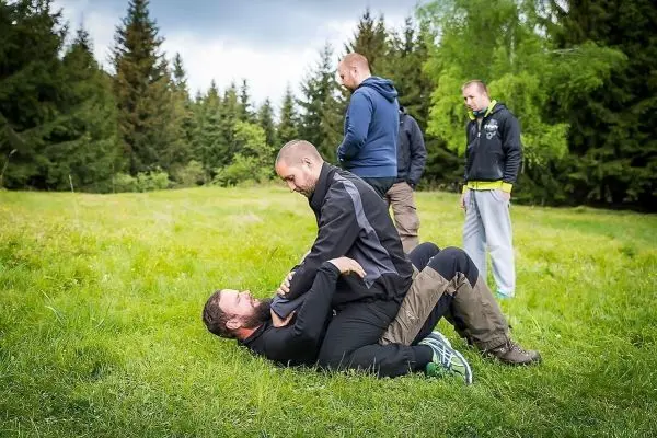 Zwei Personen üben eine Bodenkampftechnik auf einer Wiese, während andere zuschauen