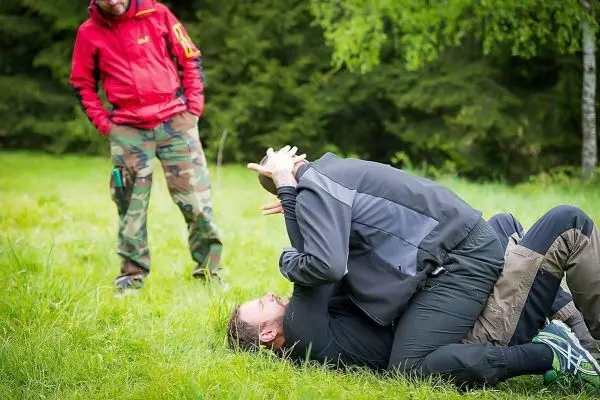 Zwei Personen üben eine Kampfsporttechnik auf einer Wiese, während eine dritte Person zuschaut